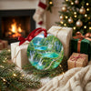 Scenic Croatian lake ornament resting near wrapped gifts and a glowing holiday fireplace.