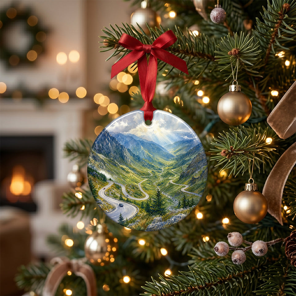 Ceramic mountain road ornament hanging from a festive Christmas tree branch surrounded by warm lights.